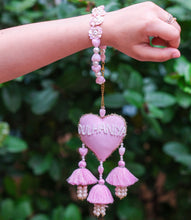 A pink heart-shaped beaded ornament with tassels and rhinestone embellishments, hanging from a bracelet worn on a person's wrist against a green leaf background.